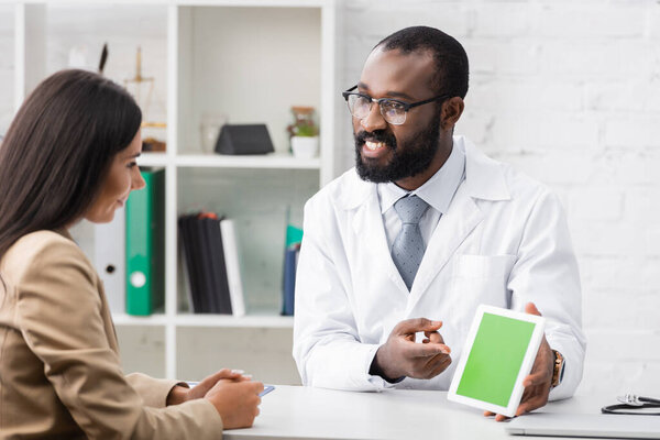 bearded african american doctor in eyeglasses pointing with finger at digital tablet with green screen near brunette patient