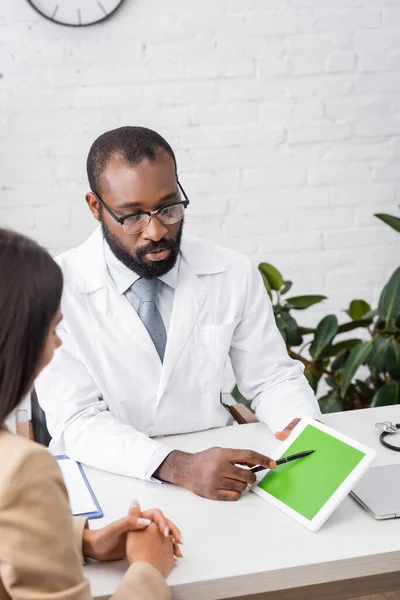 ブルネット女性の近くに緑の画面とデジタルタブレットでペンを指して眼鏡で深刻なアフリカ系アメリカ人医師の選択的焦点 — ストック写真