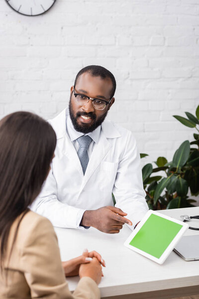 selective focus of bearded african american doctor in eyeglasses pointing with finger at digital tablet with green screen near brunette woman