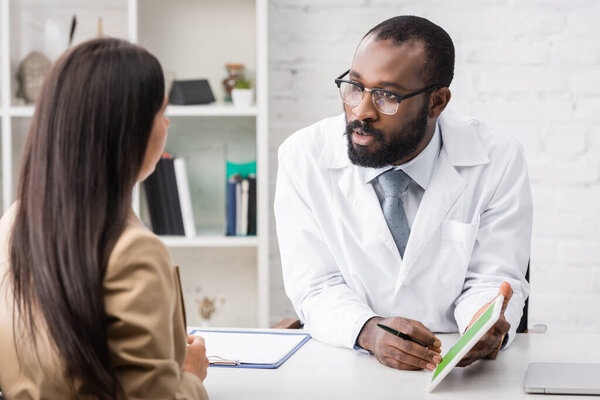 selective focus of serious african american doctor showing digital tablet while talking to brunette woman