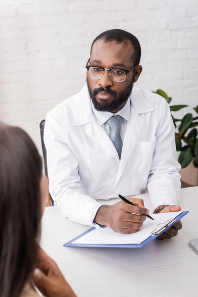 african american doctor in eyeglasses holding clipboard and pen near sick patient touching sore throat