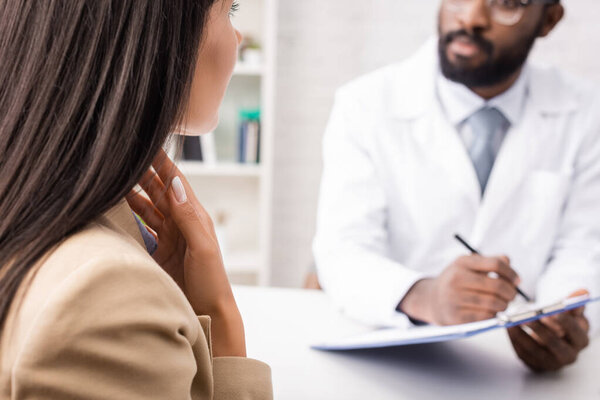 selective focus of diseased woman touching sore throat near african american doctor holding clipboard