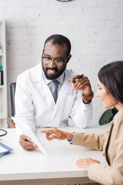 selective focus of brunette woman pointing with finger at digital tablet near african american doctor