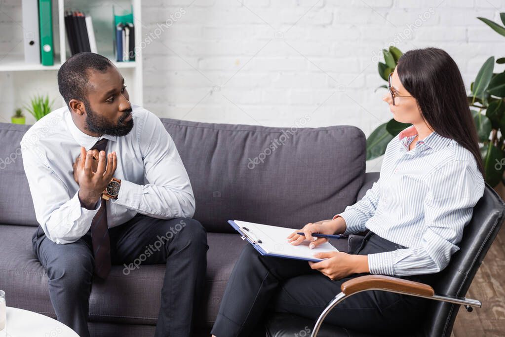 selective focus of worried african american man gesturing while talking to brunette psychologist holding clipboard