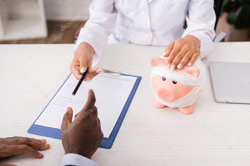 cropped view of doctor touching piggy bank, wrapped in bandage, and giving pen to african american patient near insurance claim form