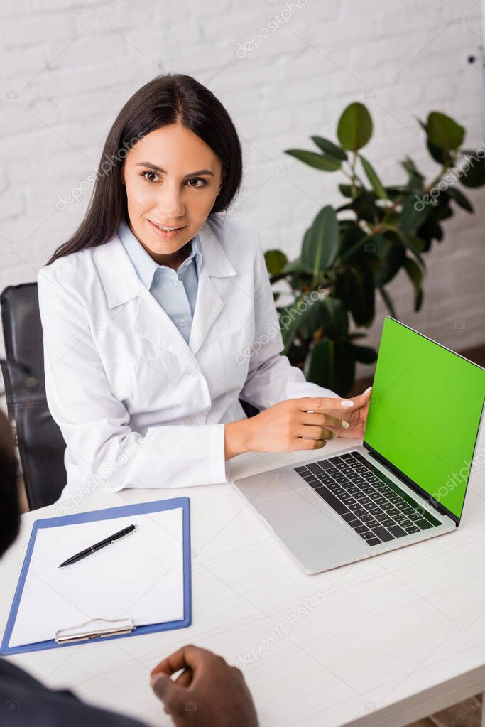 selective focus of doctor pointing with finger at laptop with green screen near african american patient 