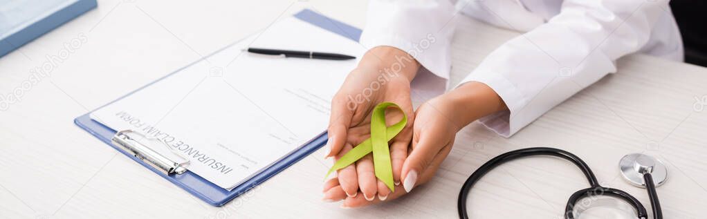cropped view of doctor holding green awareness ribbon near stethoscope and insurance claim form, mental health concept, horizontal image