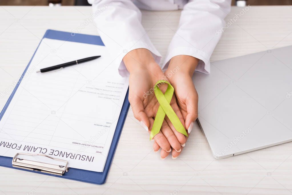 cropped view of psychologist holding green awareness ribbon near insurance claim form and laptop, mental health concept
