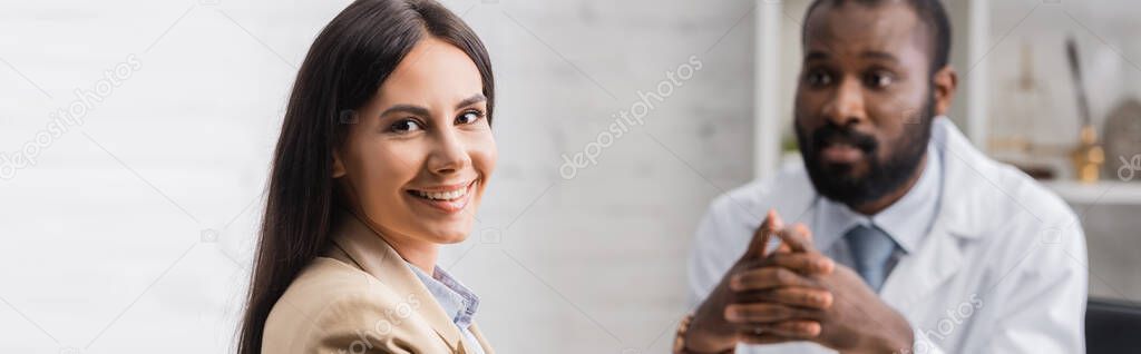 panoramic shot of joyful brunette woman looking at camera near african american doctor