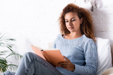 Yatak odasında süveter giyen, kitap okuyan kızıl saçlı kadının seçici odağı. 