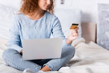 Yatak odasında online alışveriş yaparken dizüstü bilgisayar ve kredi kartı kullanan genç bir kadın görüntüsü. 