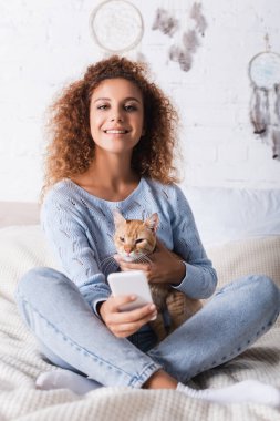 Kedi besleyen ve yatakta akıllı telefon tutan kadınların seçici odak noktası. 