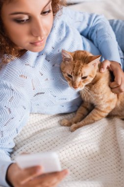 Yatakta akıllı telefon kullanırken kedi besleyen kadının seçici odağı 