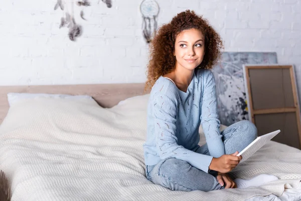 Red haired woman in sweater and jeans holding digital tablet on bed