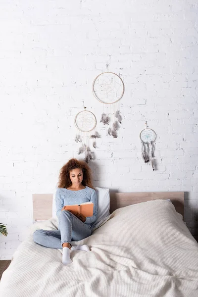 Red haired woman reading book on bed near dreamcatchers on wall