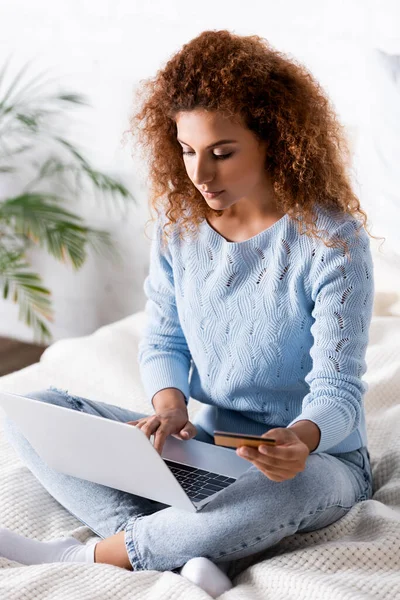 穿着牛仔裤和毛衣的女性在床上使用信用卡和笔记本电脑的选择性焦点 — 图库照片