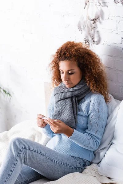 Curly woman in scarf and sweater holding thermometer on bed