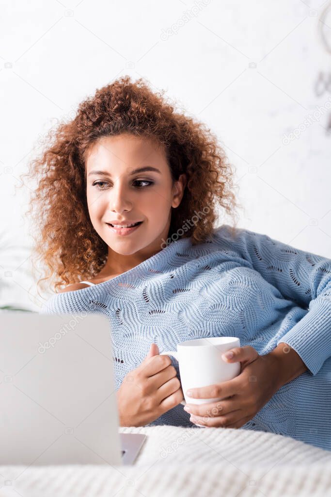 Selective focus of red haired woman in sweater holding cup near laptop on bed 