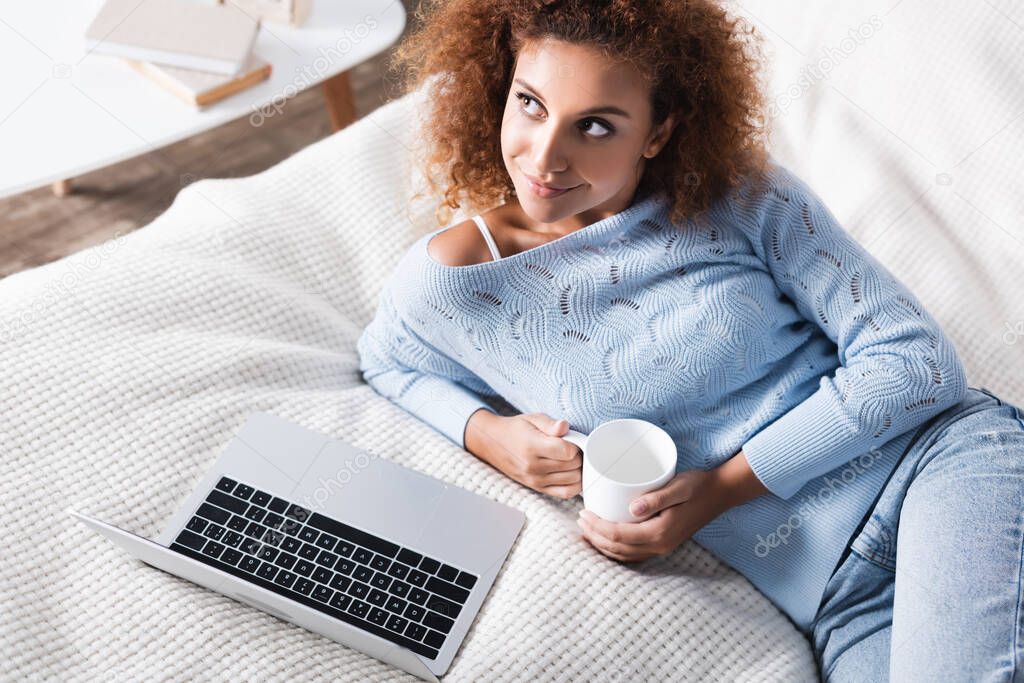 High angle view of woman holding cup near laptop on bed 