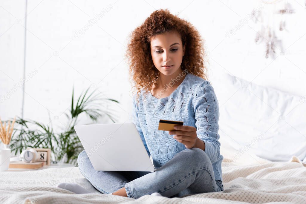 Selective focus of curly woman using laptop and credit card on bed 