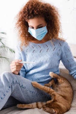 Selective focus of young woman in medical mask looking at thermometer beside tabby cat on bed   clipart