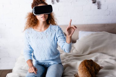 VR kulaklıklı genç kadın yatak odasındaki kedinin yanında parmağıyla işaret ediyor. 