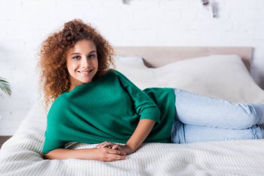 Genç kıvırcık kadın evde yatarken kameraya bakıyor. 