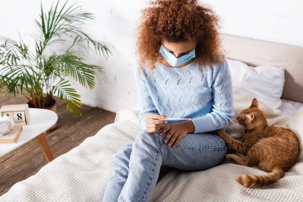 ベッドの上で猫の近くに座っている間 医療用マスク保持温度計の病気の女性 — ストック写真