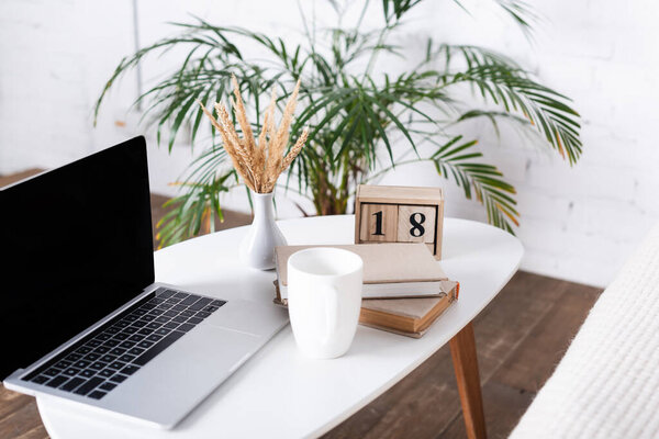 Selective focus of laptop with blank screen, books and cup on coffee table 