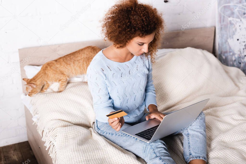 Selective focus of young woman holding credit card and using laptop near tabby cat on bed 