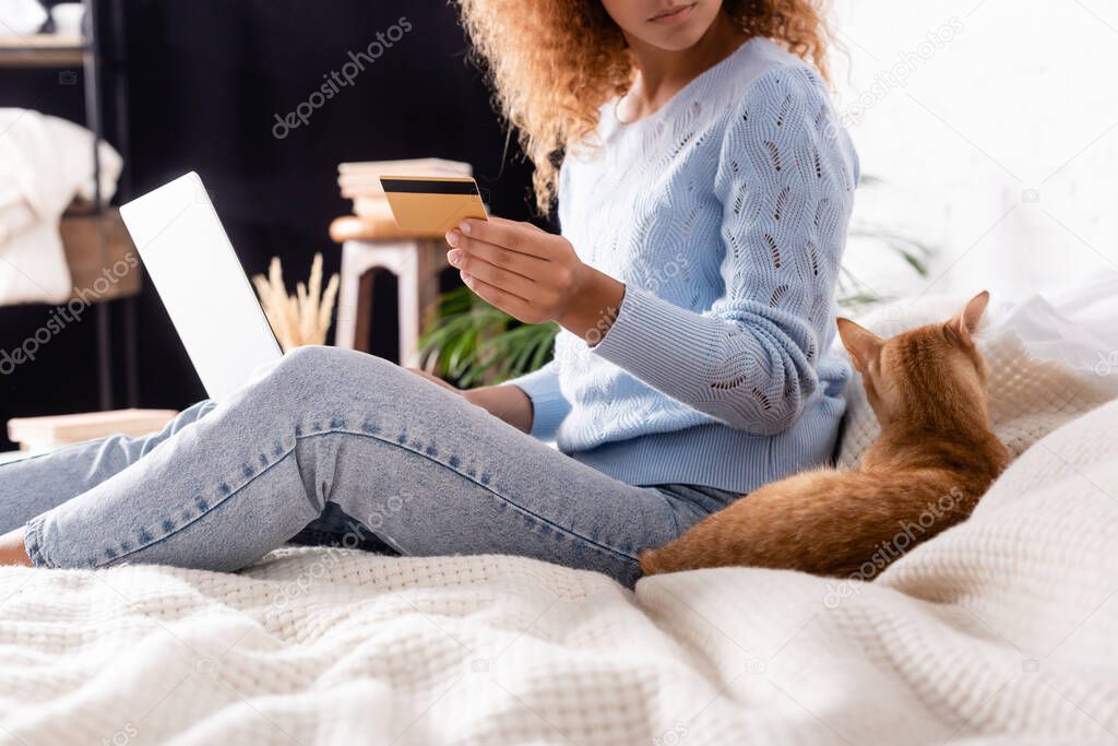 Cropped view of cat lying near woman using laptop and credit card in bedroom 