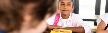 horizontal crop of african american schoolgirl sitting in school dining room near lunch box clipart