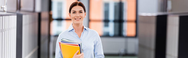 horizontal image of teacher looking at camera while standing in school corridor with folders