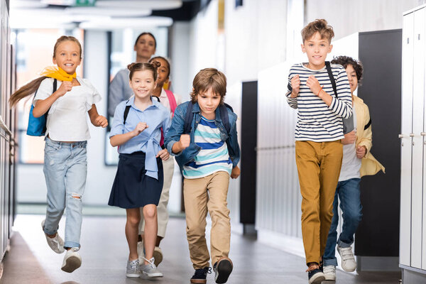 excited multicultural pupils running along school corridor with teacher on background