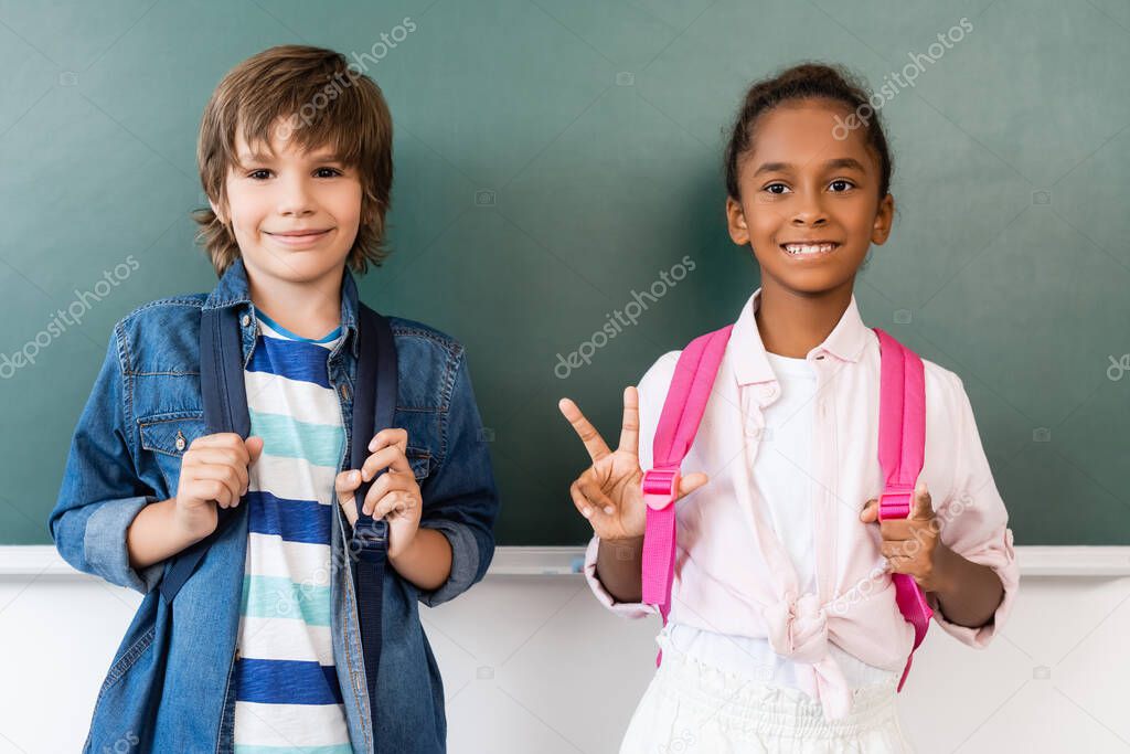 african american schoolgirl showing victory gesture while standing at chalkboard with classmate