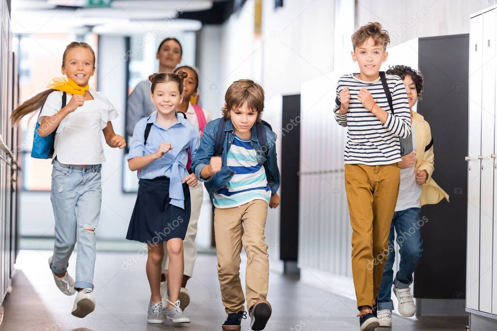 excited multicultural pupils running along school corridor with teacher on background