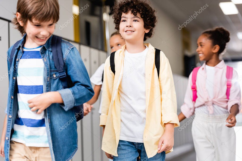excited multicultural schoolchildren walking along school corridor