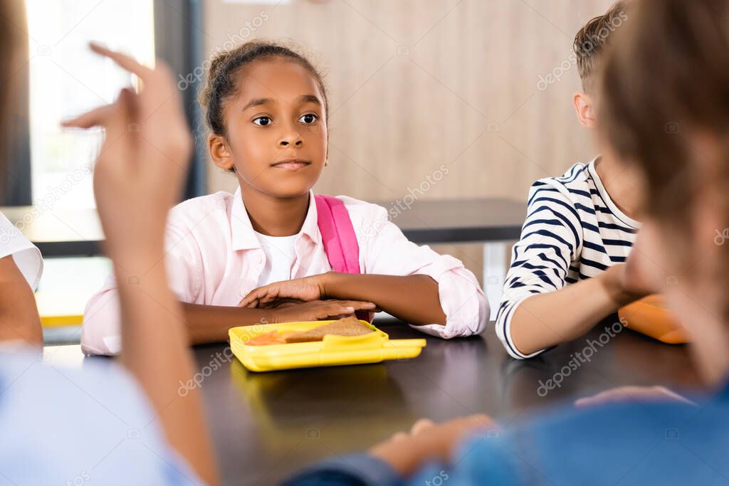selective focus of african american schoolgirl sitting at lunch box near classmates in school canteen