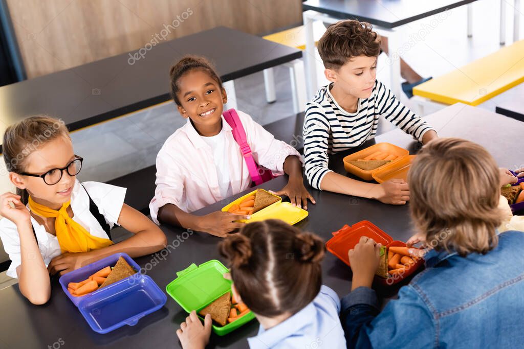 selective focus of african american schoolgirl looking at camera while sitting in school eatery near multicultural classmates