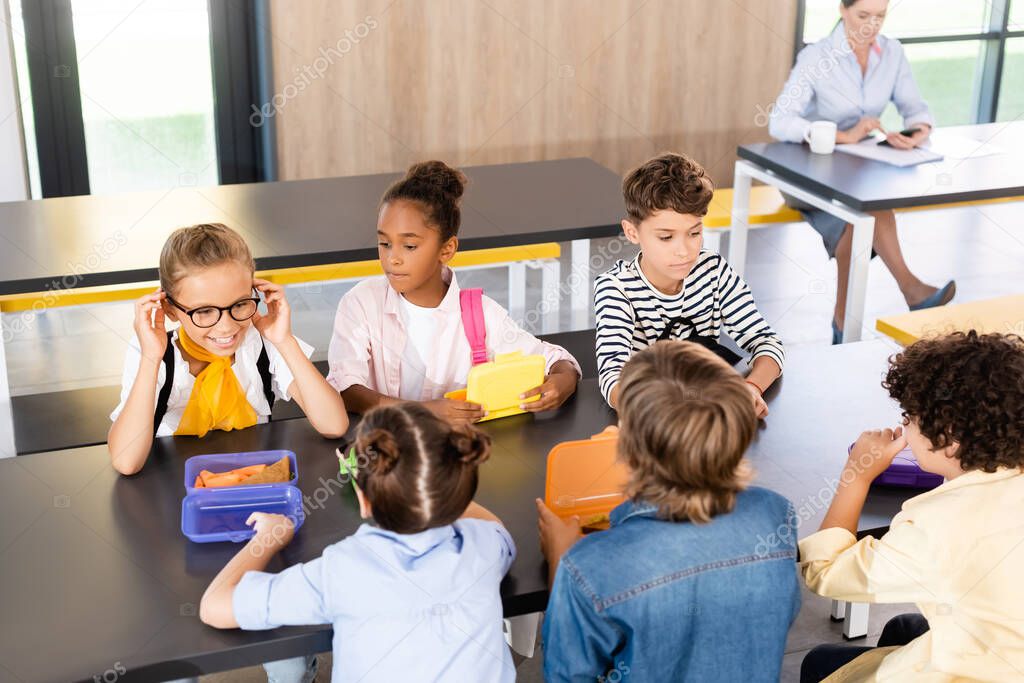 multiethnic classmates sitting in school canteen near lunch boxes and teacher on background