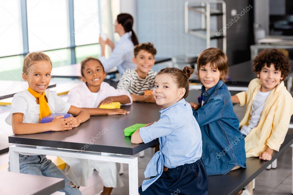 multicultural pupils looking at camera while sitting in dining room with teacher on background