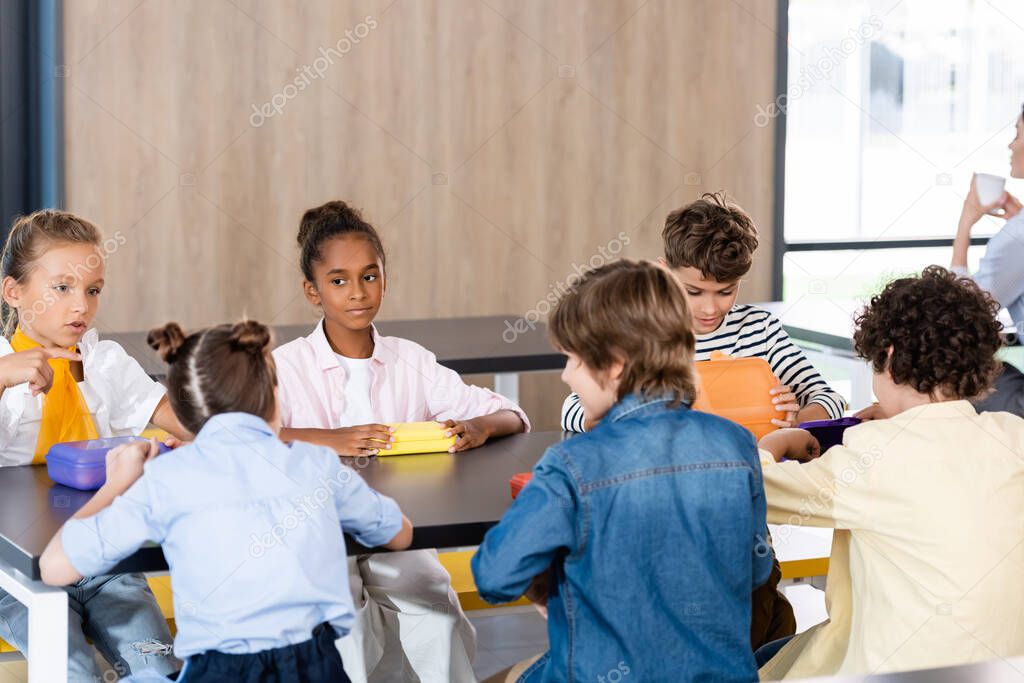 schoolgirl pointing with finger while sitting with multicultural classmates in school dining room