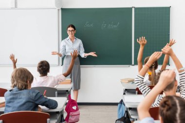back view of multiethnic pupils with hands in air, and teacher standing with open arms near chalkboard with back to school lettering clipart