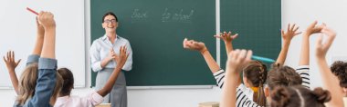 Elleri havada çok kültürlü öğrencilerin arka planı ve okula geri dönen yazı tahtasının başında duran öğretmen.
