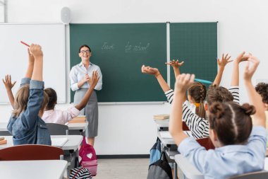 Elleri havada çok kültürlü öğrencilerin arka planı ve okula geri dönen yazı tahtasının başında duran öğretmen.