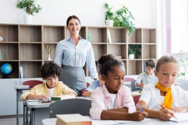 Selective focus of teacher looking at camera near multiethnic pupils during lesson in school   clipart