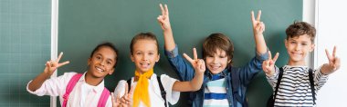 Horizontal crop of multiethnic classmates showing peace sign at camera near green chalkboard  clipart