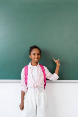 African american schoolgirl pointing with finger at chalkboard in classroom  clipart