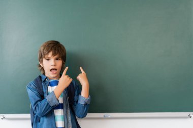 Sırt çantalı heyecanlı öğrenci sınıftaki yeşil karatahtayı işaret ediyor. 