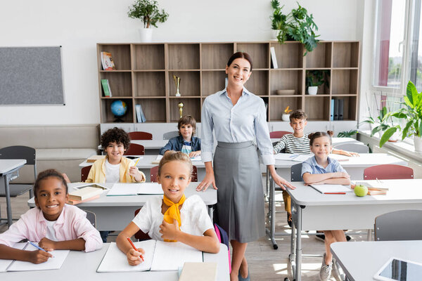 teacher and multiethnic pupils looking at camera in classroom during lesson